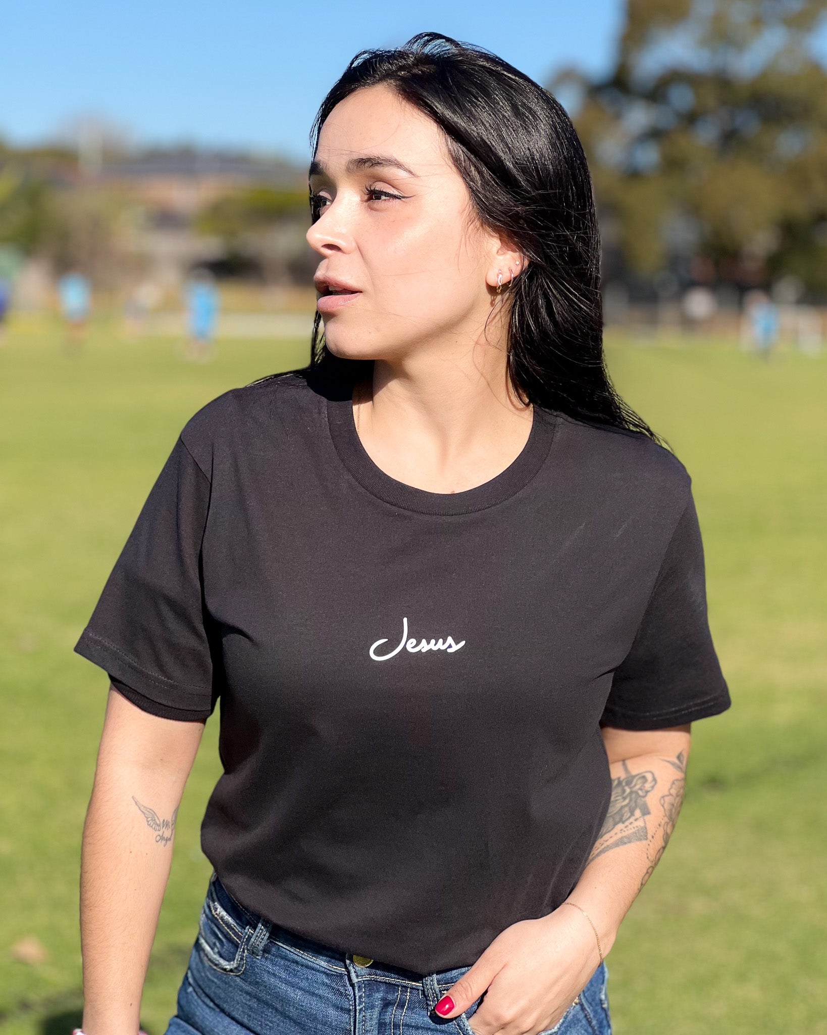 A woman standing in an open field wearing a christian black crew neck t-shirt with the text 'Jesus' written on the front in a white cursive font.