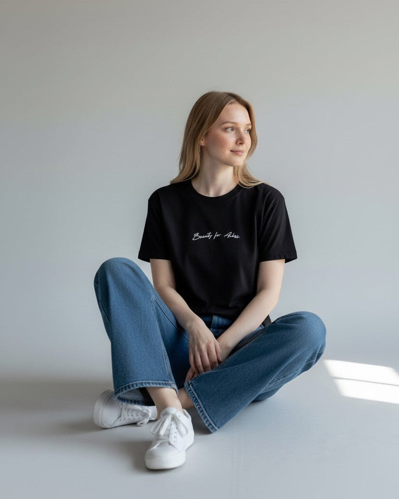 Person wearing a black t-shirt and blue jeans sitting on the floor against a plain wall.
