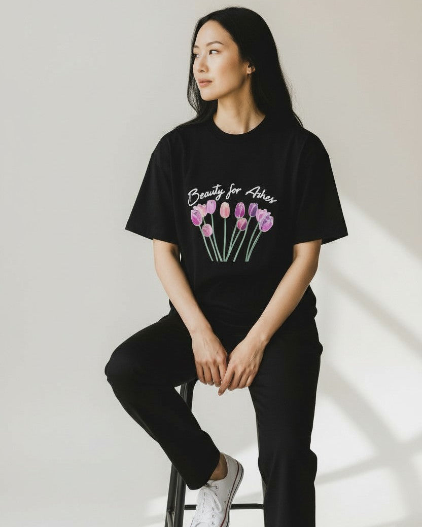 Person wearing a christian black t-shirt with floral design and text, sitting on a stool against a white wall.