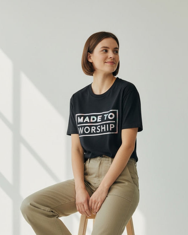 Woman sitting on a stool wearing a black t-shirt with 'Made to Worship' text in a minimalistic setting.