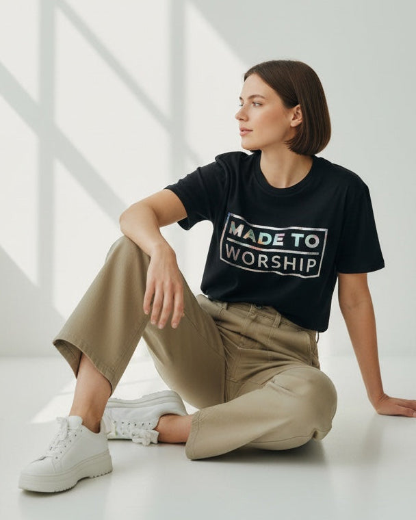 Woman wearing a black t-shirt with 'Made to Worship' text, sitting on a white floor with sunlight streaming through a window.