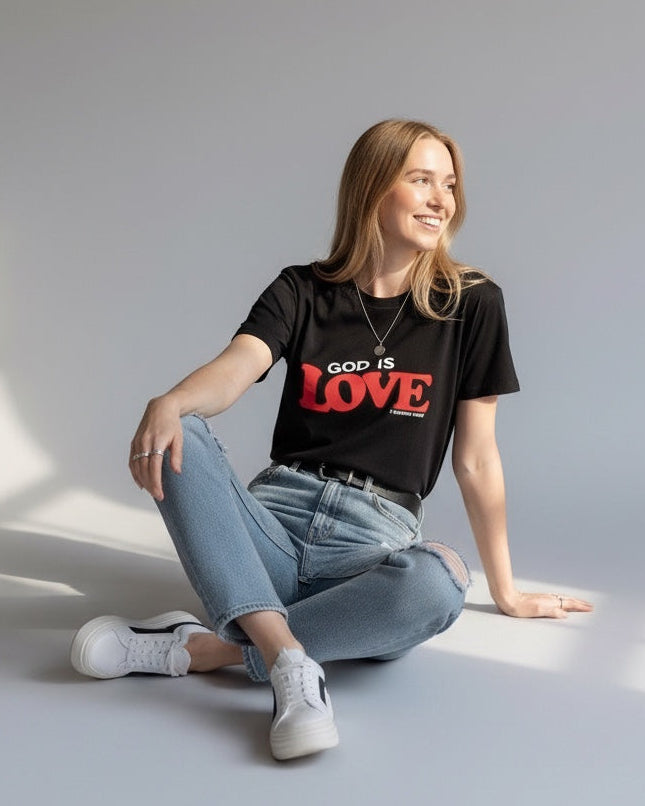 Woman wearing a black t-shirt with 'God is Love' text, sitting on the floor with a neutral background